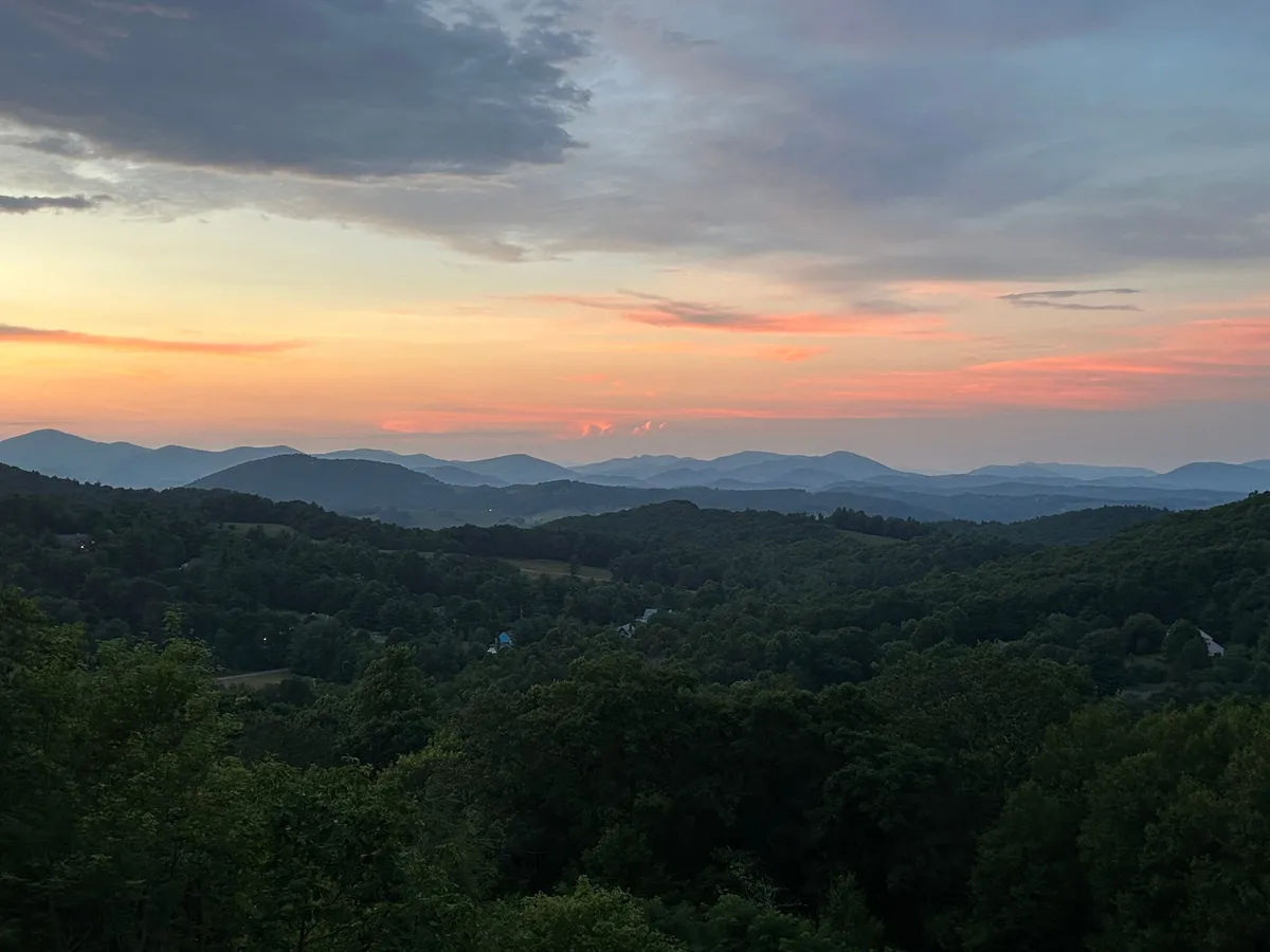 A sweeping view of the Blue Ridge Mountains, with verdant green valleys stretching into the distance and a sky ablaze with the colors of sunset. The expansive landscape encapsulates the natural beauty and awe-inspiring vistas that draw visitors to the Parkway.