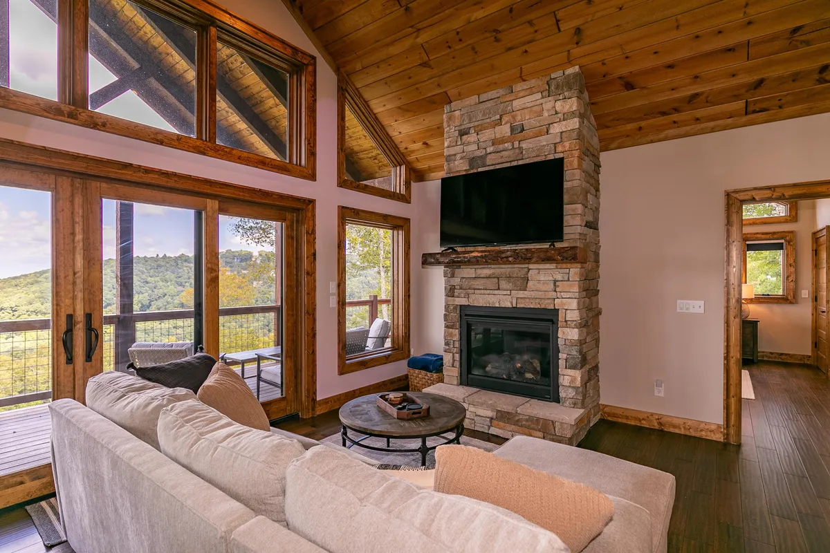 A spacious living room featuring large windows that frame sweeping mountain views. A comfortable sofa faces a stone fireplace, creating a cozy setting. The round coffee table and warm lighting accentuate the inviting atmosphere, and the natural landscape visible through the windows draws the eye outward.