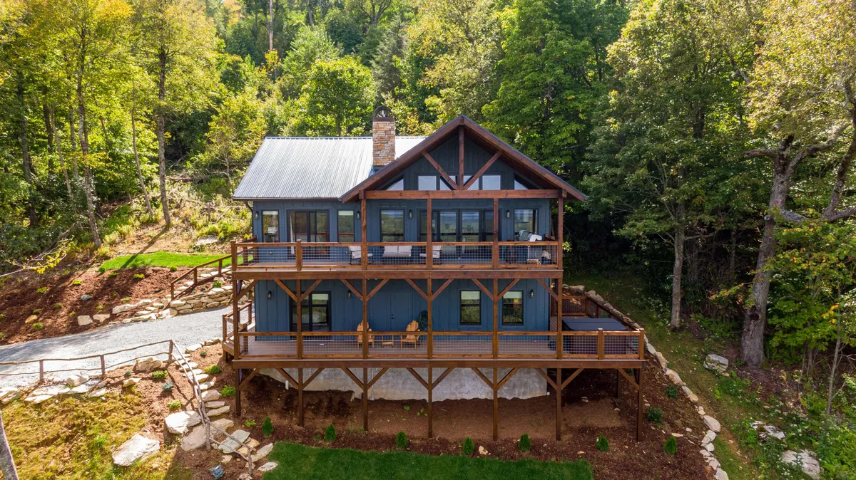 An inviting wooden deck wraps around the house, framed by lush greenery and large windows reflecting the natural surroundings. The stone chimney adds rustic charm, while the open deck provides a perfect vantage point for watching sunsets or relaxing after an exploration-filled day.