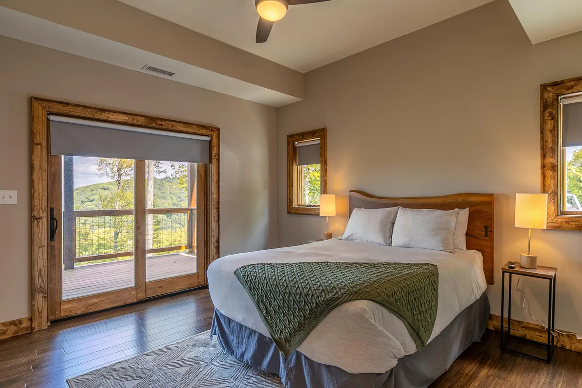A serene bedroom with a green throw blanket on the bed, wooden framed windows, and a lamp-lit nightstand. The ample windows suggest morning light and views of the surrounding mountains, reinforcing the connection to the outdoors.