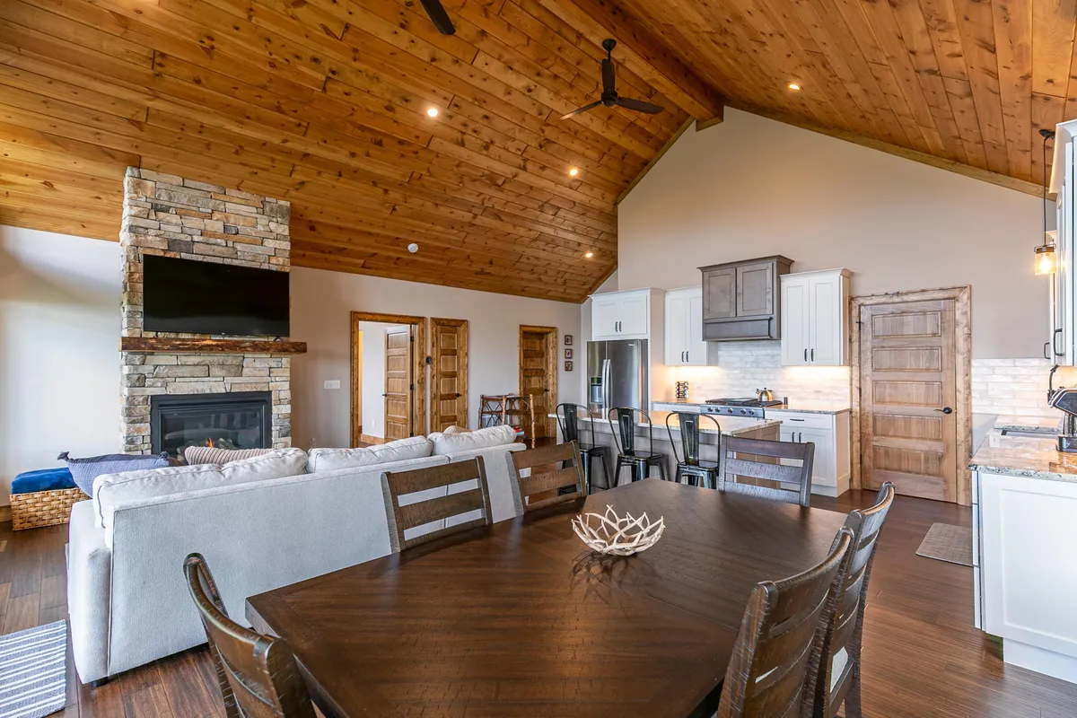 A cozy dining and kitchen space featuring a stone fireplace, wooden beams, a dining table, and a kitchen island. The setting is ideal for group meals and relaxing by the fire after outdoor activities.