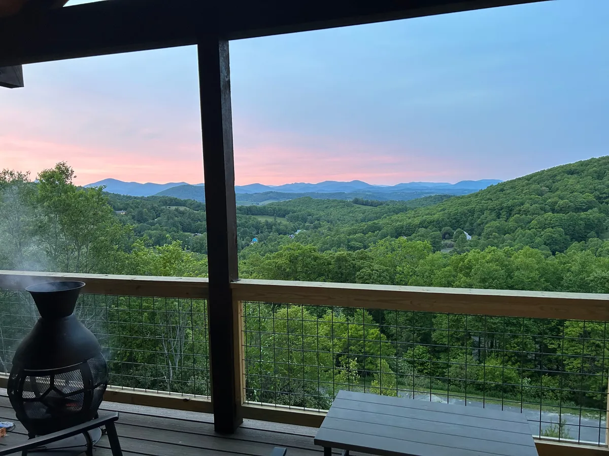 The image features an inviting deck scene with outdoor furniture and a rustic chiminea in the foreground, set against a backdrop of vibrant sunset hues and mountain vistas. The atmosphere suggests warmth and tranquility, perfect for sipping wine and unwinding by the fire.