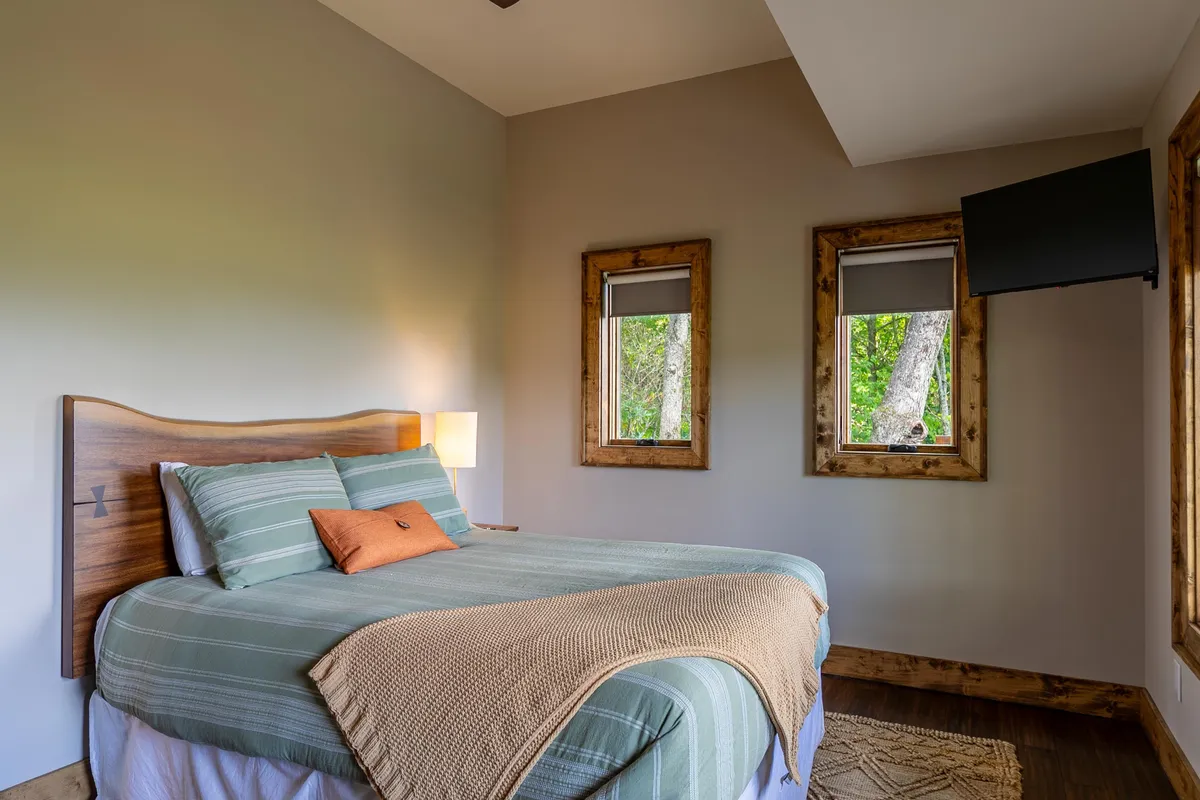 A cozy bedroom with a plush bed, windows welcoming natural light, a lamp for cozy evenings, and a television for personal entertainment. The setup feels inviting, offering a sense of warmth and privacy.