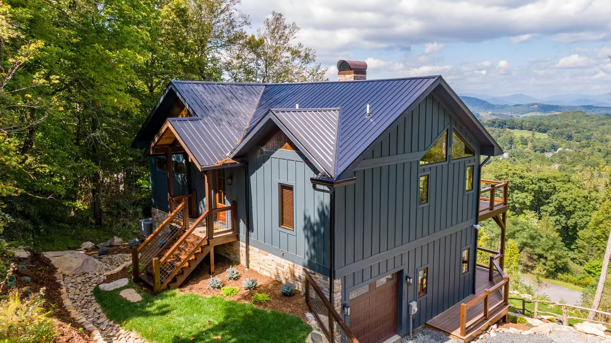 The hero image showcases the striking blue exterior of a custom-built house set amidst lush trees, with expansive mountain vistas in the background. Wooden stairs lead up to an inviting deck, perfect for outdoor dining or watching sunsets. The serene setting hints at nearby hiking trails and a peaceful escape in nature.