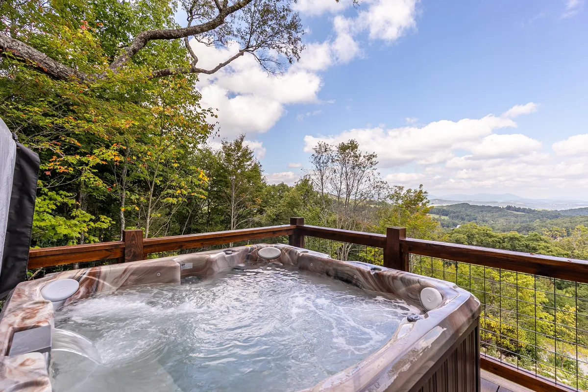 This exterior image features a hot tub on the lower deck, bordered by a wooden railing and overlooking majestic long-range views of the Blue Ridge mountains. The serene setting invites relaxation, with hints of sunset lighting and opportunities for stargazing or early morning tranquility.