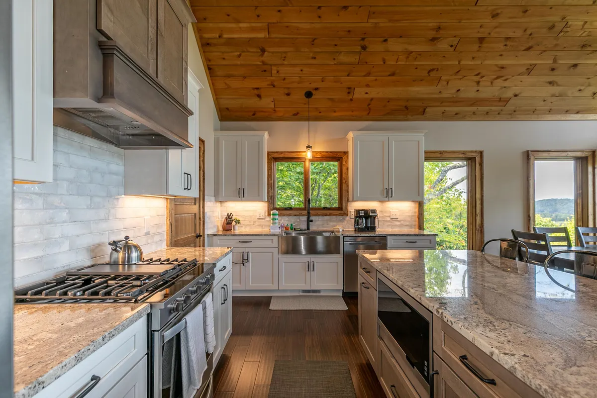 A modern kitchen with granite countertops, stainless steel appliances, a wooden ceiling, and large windows providing scenic views. The kitchen exudes warmth and is well-equipped for preparing meals and hosting family events.