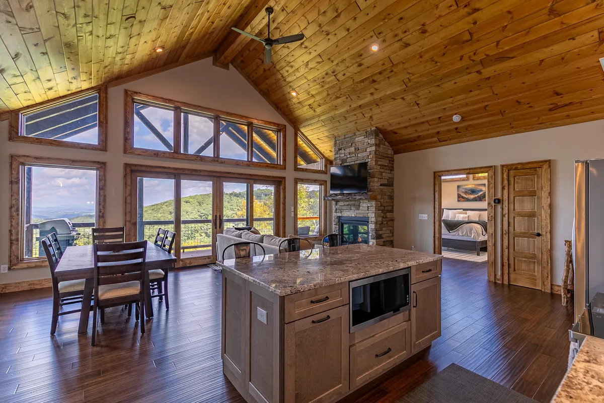 An open kitchen and dining space with a granite island, wooden dining table, and expansive windows that showcase beautiful mountain scenery. The area is filled with natural light, inviting relaxation and togetherness while keeping the landscape as a central feature.