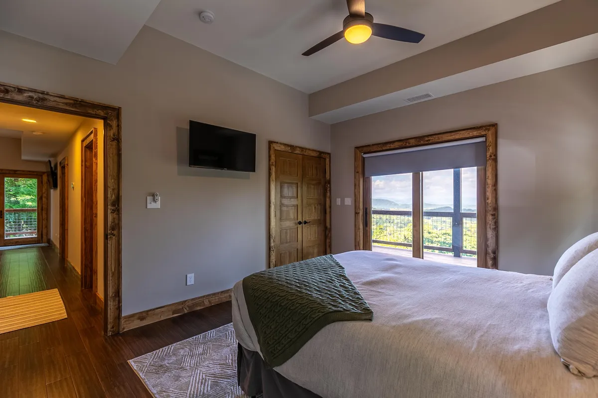 This image features a cozy bed with a green throw, a large window that frames an inviting view of the outdoors, and warm wooden accents. The broad window draws the eye outward, suggesting the mountain scenery and seasonal changes guests can enjoy from the comfort of the interior.
