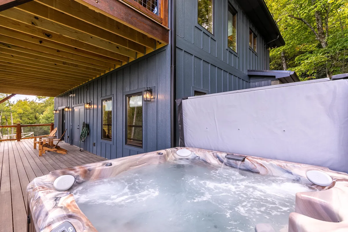 An outdoor scene highlighting the hot tub on a deck, complemented by wooden chairs and the exterior wall of the house. The image suggests a peaceful space for unwinding, socializing, or enjoying nature's sounds, with the backdrop of sunset or starlit sky possibilities.
