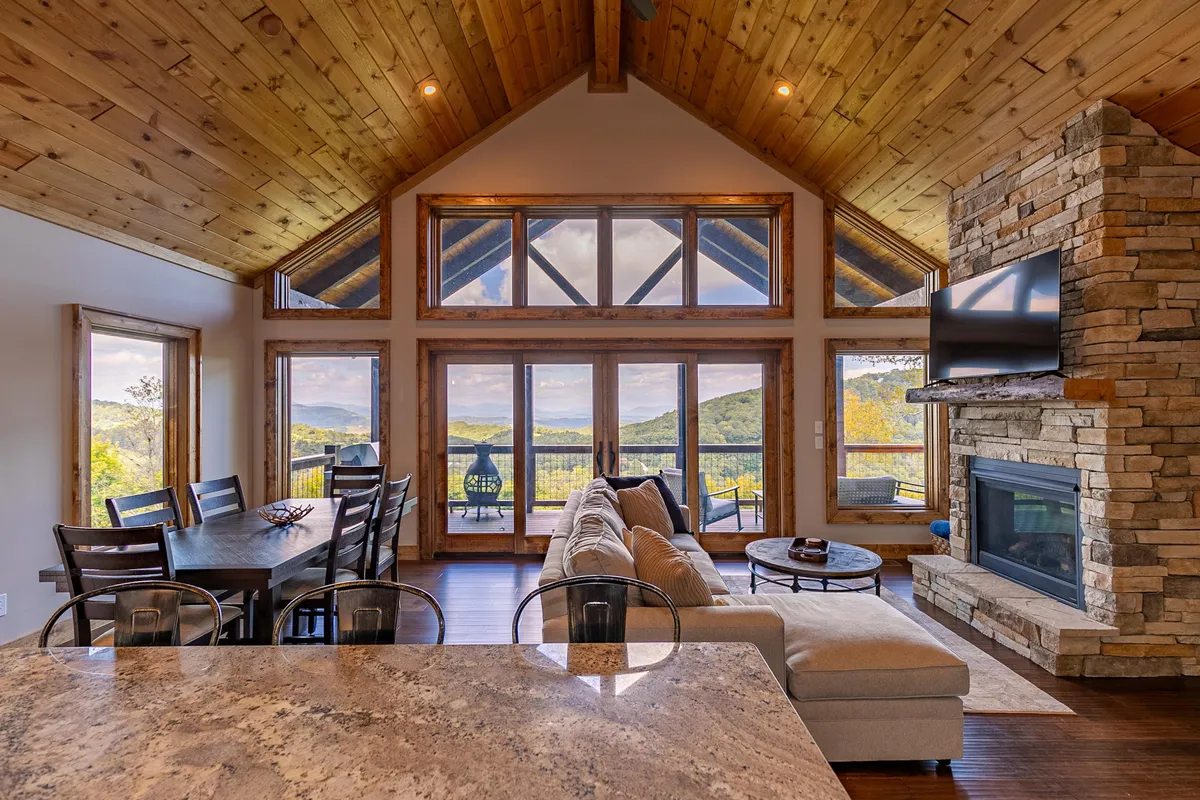 The image showcases the great room with expansive windows offering sweeping mountain views. A plush sectional sofa faces a striking stone fireplace, while a wooden dining table is positioned to take full advantage of the scenery. Vaulted tongue & groove ceilings soar overhead, bathing the entire space in natural light and creating an inviting, open atmosphere perfect for gatherings.