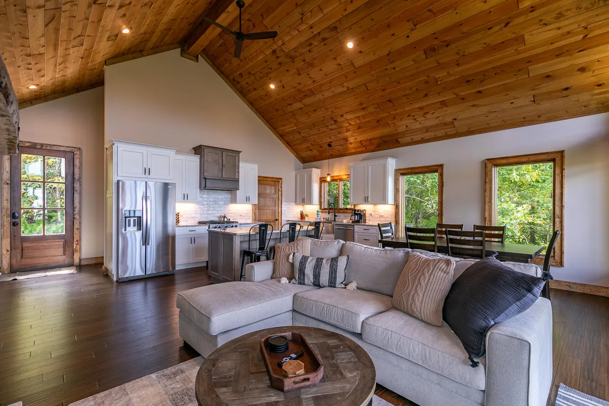 This image shows an inviting great room with a cozy sofa, a large dining table, a kitchen island, and expansive windows that let in natural light. The space is arranged for family gatherings and entertaining, and the view through the windows hints at a mountain setting.